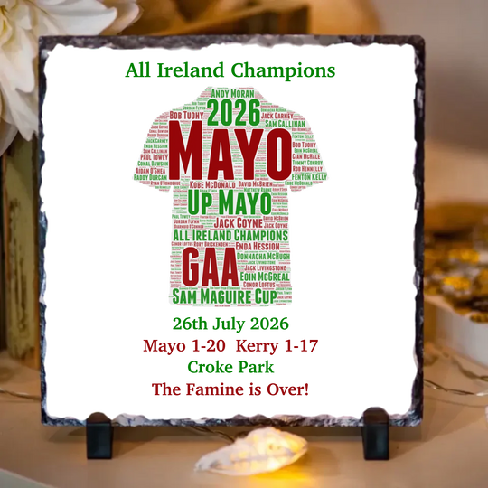 A square decorative sign on a small black stand, featuring a white background with a green and red text design celebrating Mayo GAA and the 2026 Sam Maguire Cup. The sign has a rough dark border and is photographed indoors on a tabletop with blurred decor in the background.