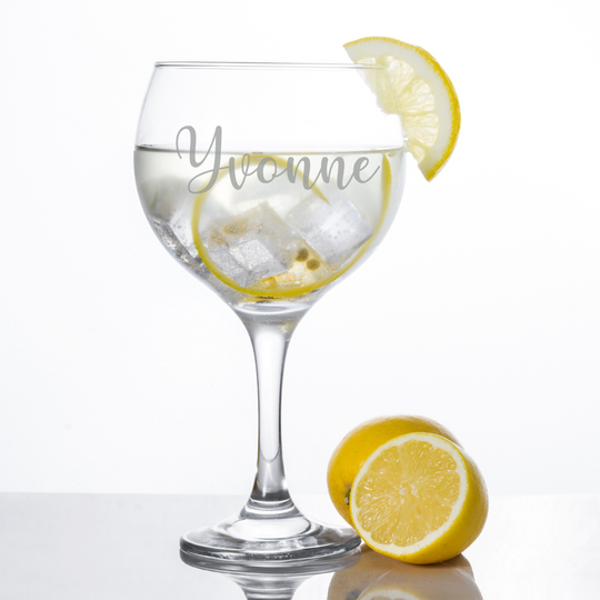 A clear glass gin glass with 'Yvonne' etched on the side, filled with a liquid and ice, accompanied by a slice of lemon. Another lemon is beside the glass on the surface.