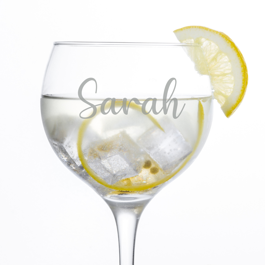 A close up of a clear glass gin glass with 'Yvonne' etched on the side, filled with a liquid and ice, accompanied by a slice of lemon. Another lemon is beside the glass on the surface