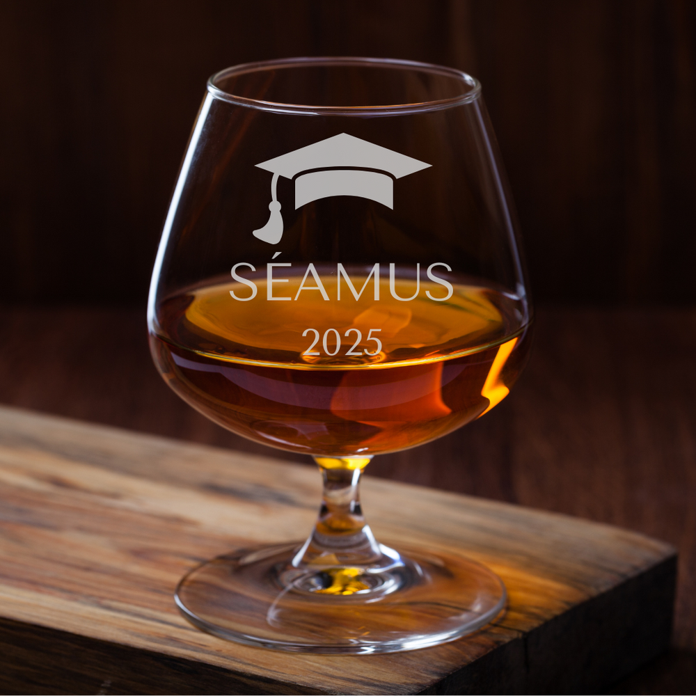 Glass filled with amber liquid, featuring a graduation cap and 'Séamus 2025' text on a wooden surface.