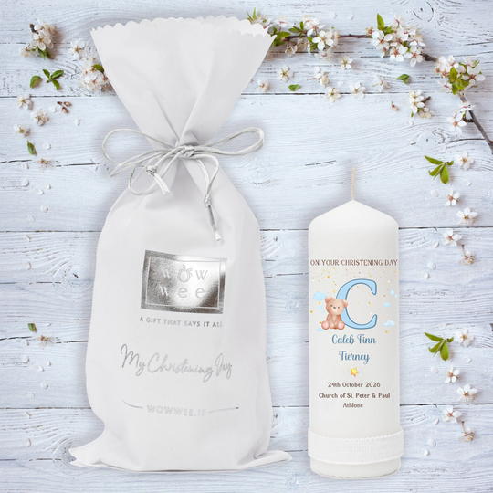 A personalized baby christeningcandle featuring a white drawstring pouch and a matching white candle on a light blue wooden background decorated with small white blossoms.