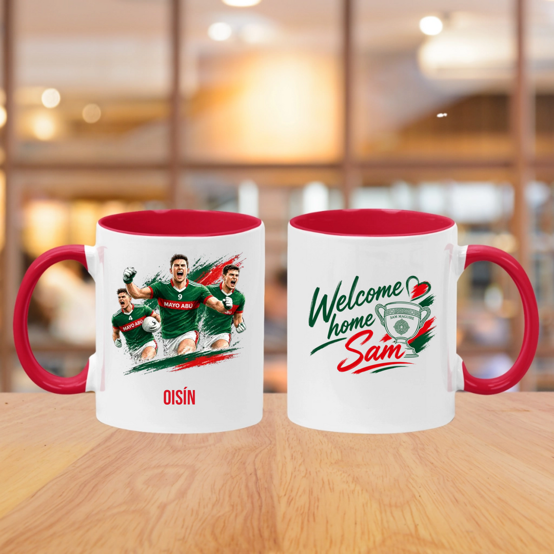 Two white ceramic mugs with red interiors and handles sit on a wooden table in a blurred indoor setting. The left mug features a printed graphic of three rugby players in green jerseys with the name 'OISÍN' below.