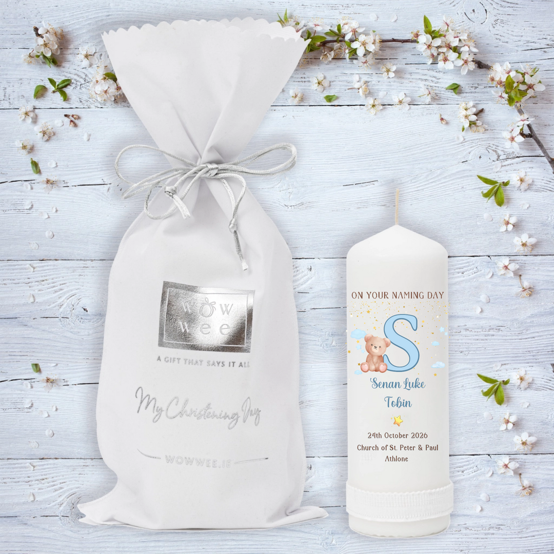 A personalized baby christening candle featuring a white drawstring gift bag and a matching white candle on a light blue wooden background decorated with white blossoms. T