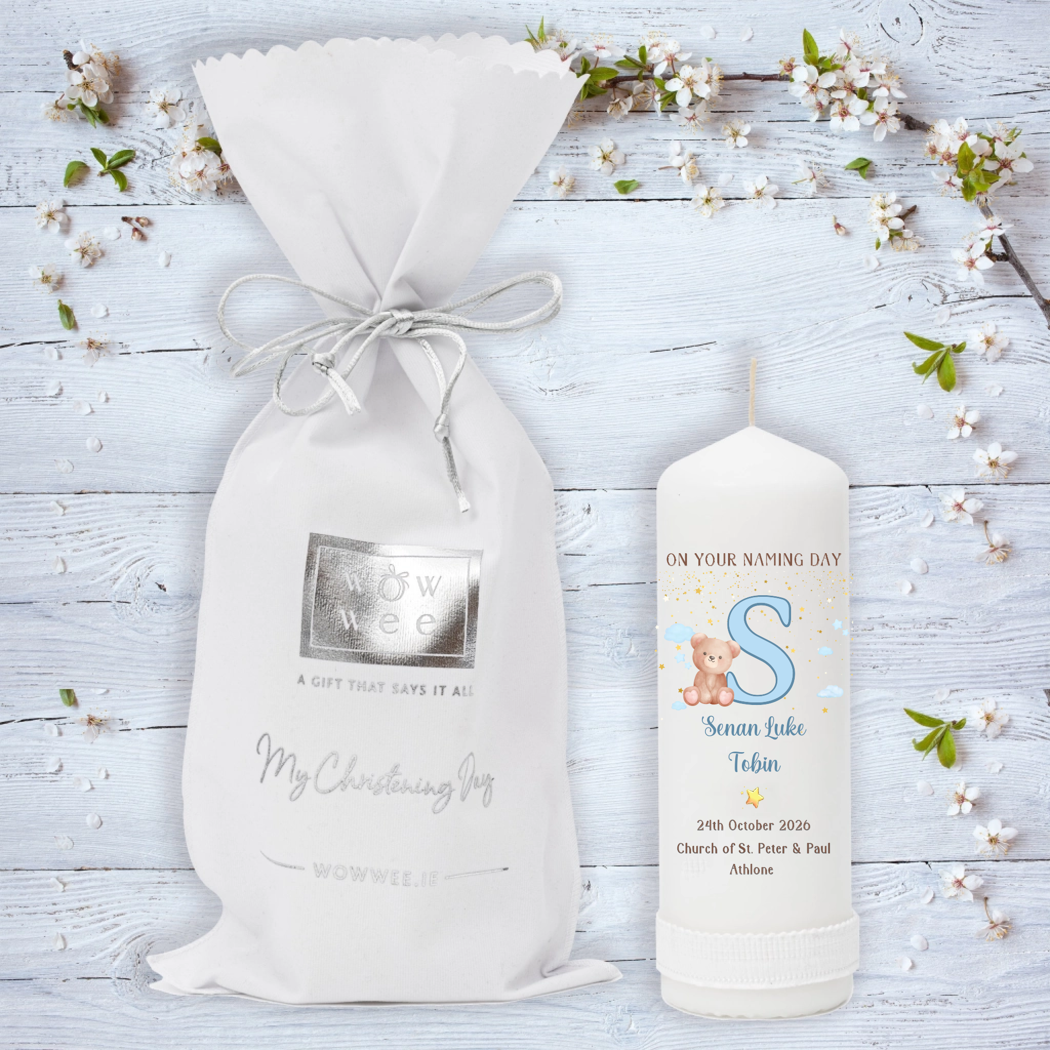 A personalized baby christening candle featuring a white drawstring gift bag and a matching white candle on a light blue wooden background decorated with white blossoms. T