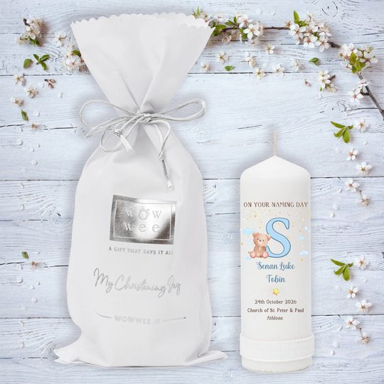 A personalized baby christening candle featuring a white drawstring gift bag and a matching white candle on a light blue wooden background decorated with white blossoms. T