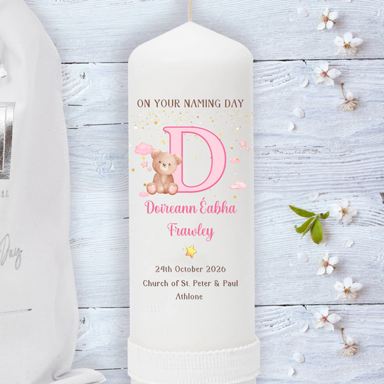 A personalized white pillar candle with a pink letter D, teddy bear illustration, and custom text for a baby naming ceremony, displayed on a light blue wooden background with decorative flowers.