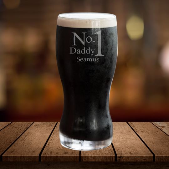 A black tulip pint glass with white foam on top, featuring a personalized name 'Seamus' etched on the side, presented on a wooden surface with a blurred background.