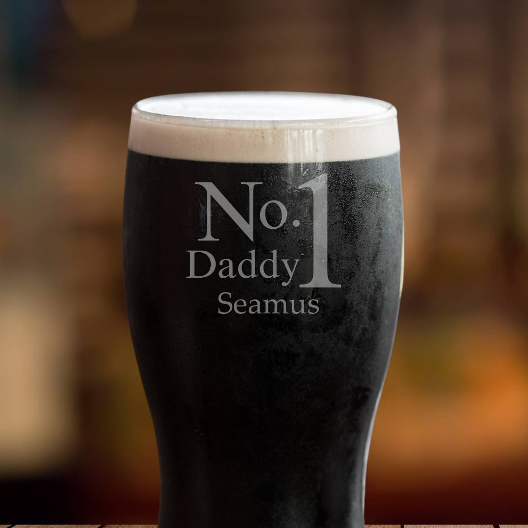 A tulip pint glass with white foam on top, featuring a personalized name 'Seamus' etched on the side, presented on a wooden surface with a blurred background.