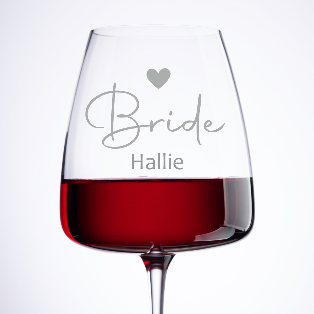 A personalized red wine glass with the word 'Bride' and a heart symbol above the name 'Hallie' etched on it, filled with red wine.