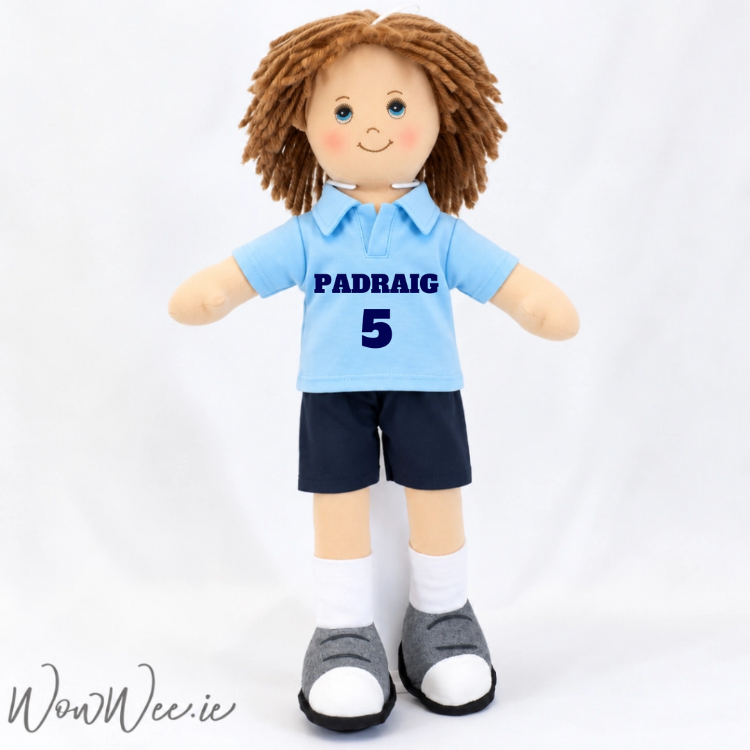 A handmade plush doll with brown yarn hair, blue eyes, and a smiling face, wearing a light blue polo shirt with 'PADRAIG 5' printed in navy, navy shorts, white socks, and grey-and-white shoes, set against a white fabric backdrop.