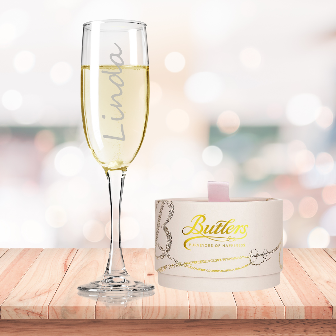 A personalized fine glass champagne flute filled with champagne, alongside a box of Butlers Powder Puff chocolates, placed on a wooden surface with a blurred background.