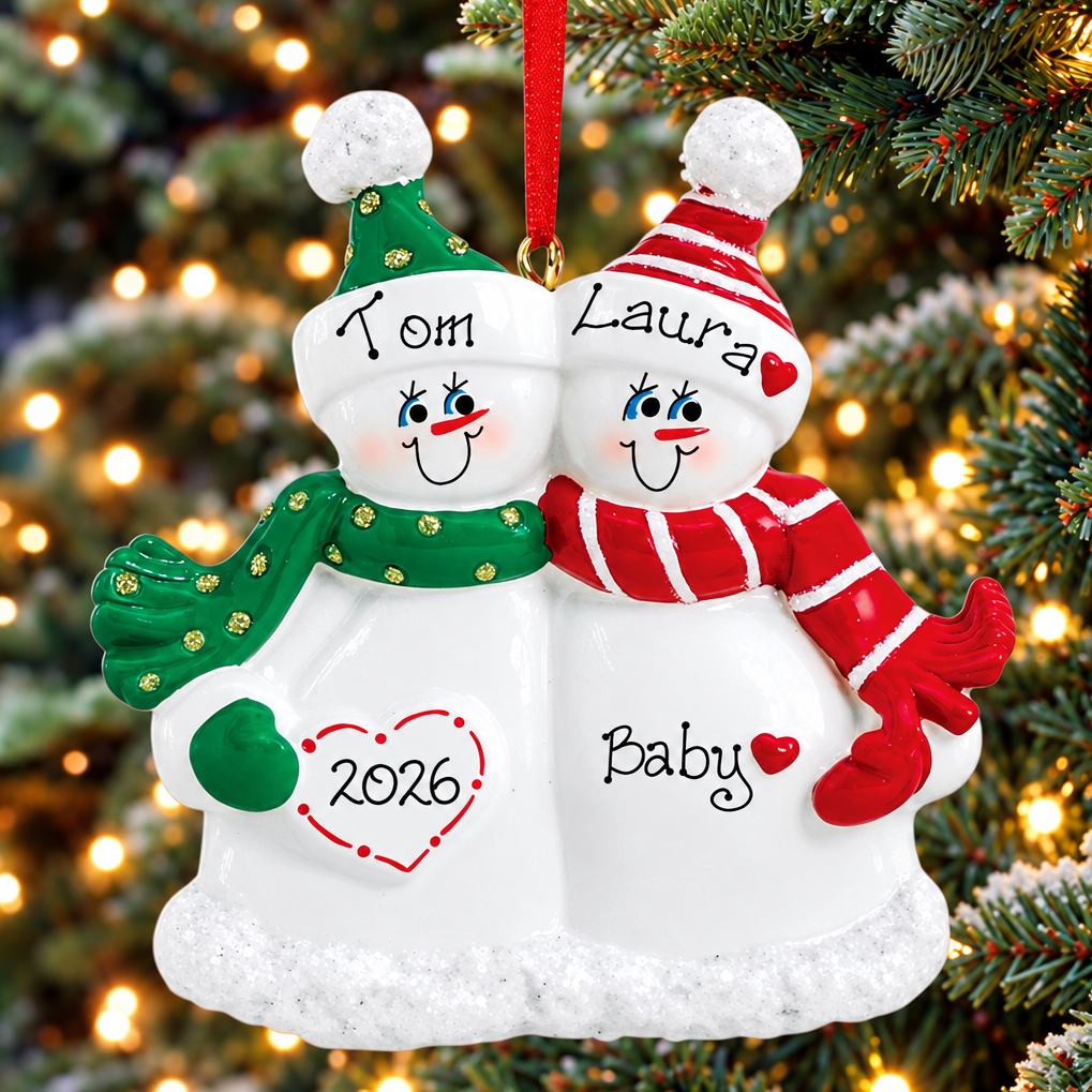 Personalized Christmas snowman ornament hanging on a Christmas tree. The ornament features two smiling snowmen side by side, one wearing a green hat and scarf with 'Tom' and '2026' on the left, and the other wearing a red and white striped hat and scarf with 'Laura' and 'Baby' on the right.