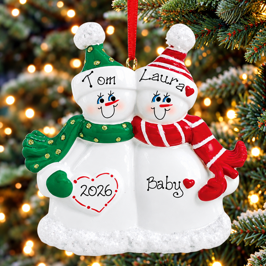 Personalized Christmas snowman ornament hanging on a Christmas tree. The ornament features two smiling snowmen side by side, one wearing a green hat and scarf with 'Tom' and '2026' on the left, and the other wearing a red and white striped hat and scarf with 'Laura' and 'Baby' on the right.