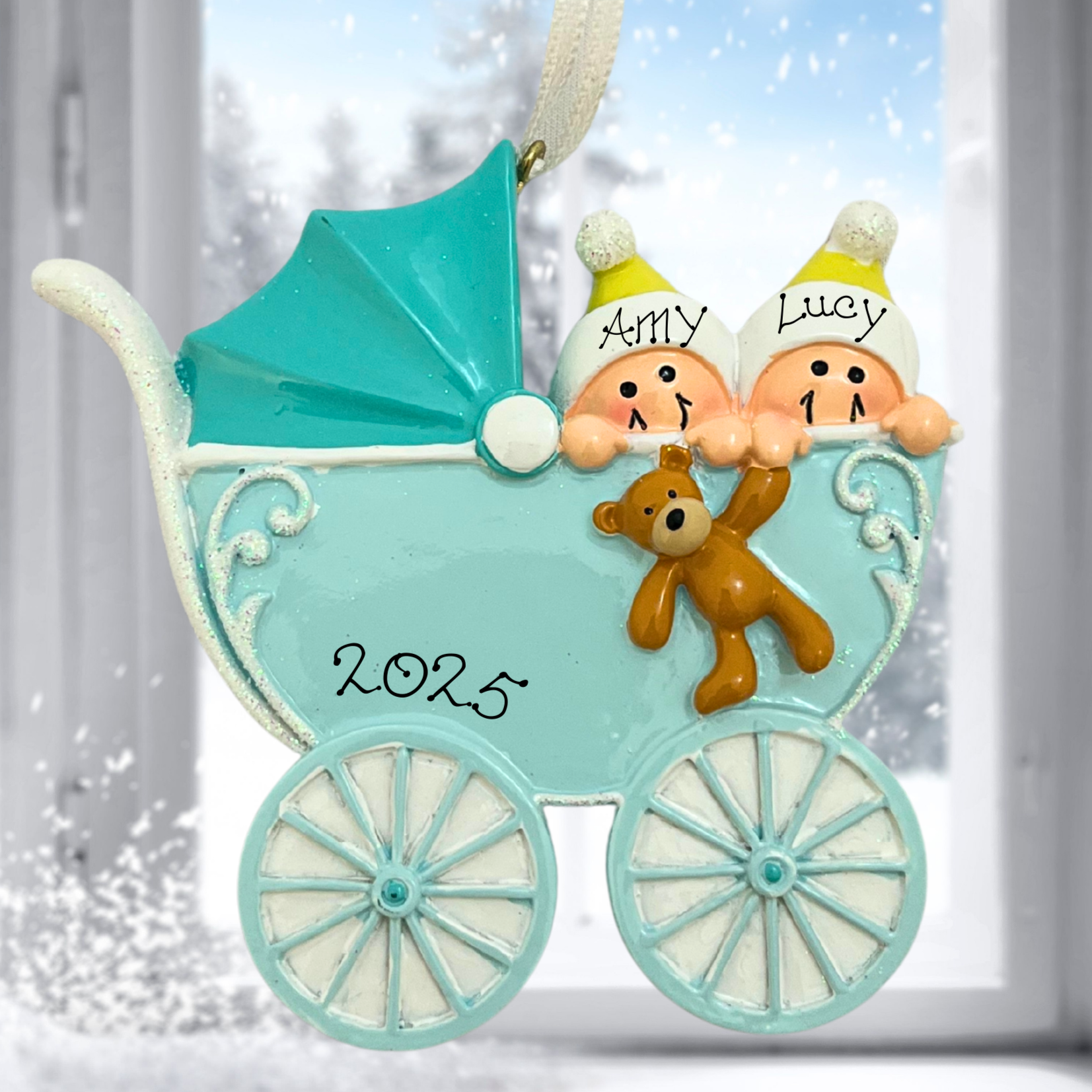 Decorative ornament of a baby carriage with two children and a teddy bear, set against a snowy window background.