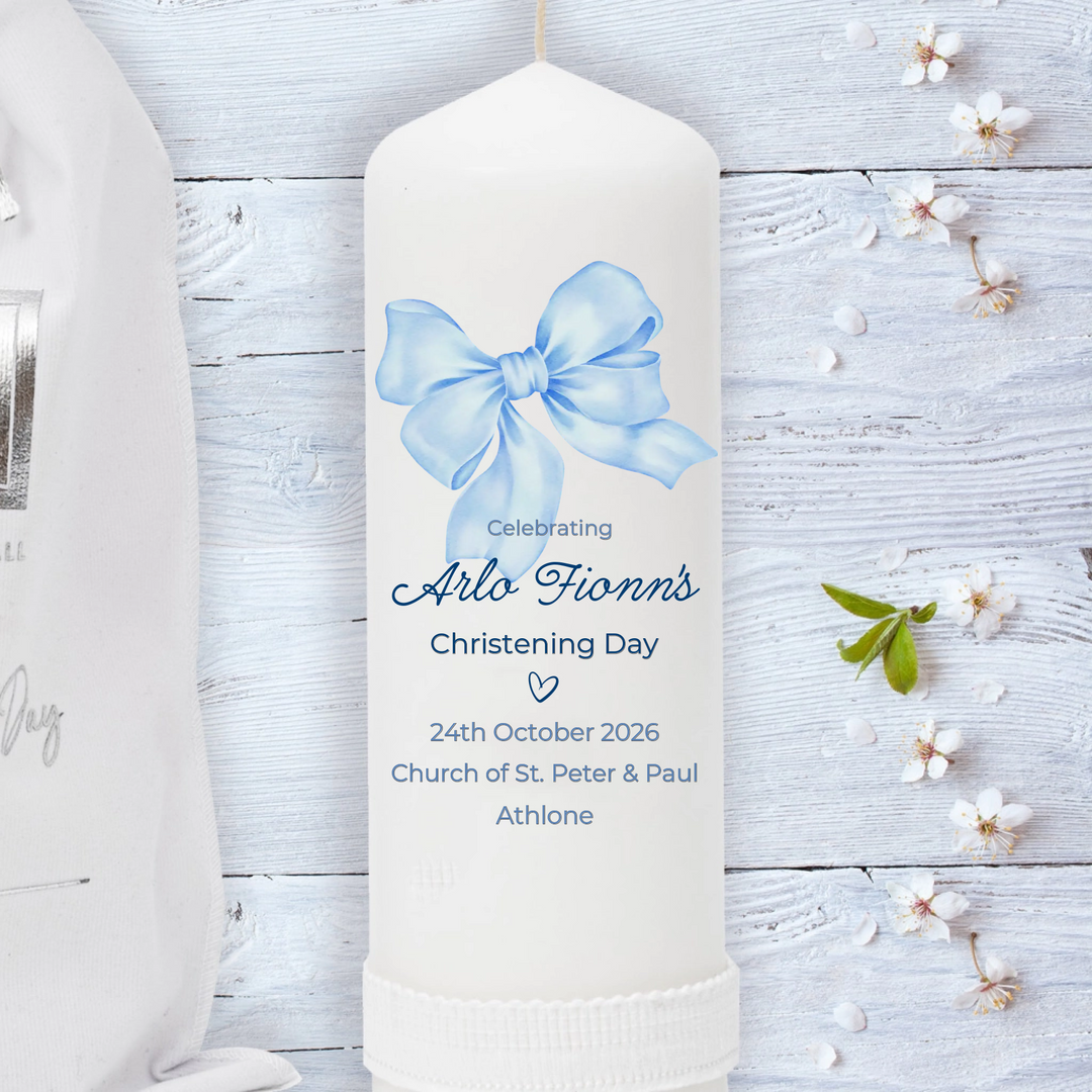 White candle with a blue bow and personalized text on a light wooden background with flowers.